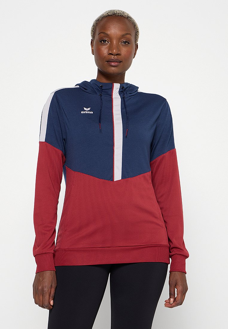 Frau in einem navy, weißen und roten farbblockierten Hoodie mit Frontreißverschluss und schwarzen Leggings, steht vor einem schlichten weißen Hintergrund.