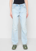 Person, die hellblaue Jeans mit geradem Bein, schwarzes Oberteil und grüne Sneakers trägt, steht vor einem schlichten grauen Hintergrund.