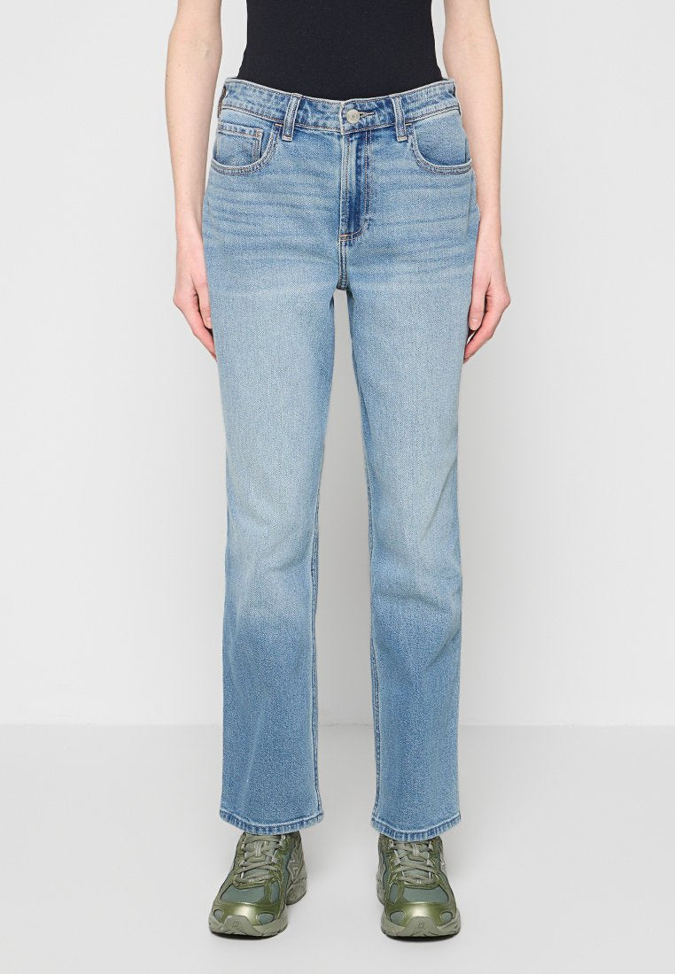 Hellblaue Jeans mit geradem Bein, getragen mit grünen Turnschuhen und einem schwarzen Oberteil, von der Taille bis zu den Füßen vor einem einfarbigen Hintergrund gezeigt.