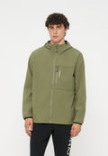 Junger Mann mit lockigem Haar, der eine olivgrüne Jacke mit Reißverschluss und Kapuze sowie schwarze Hosen mit weißem Text am Bein trägt.