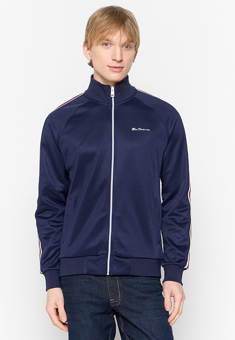 Marineblaue Zip-up Trainingsjacke aus glattem Stoff, mit weißen und roten Streifen an den Ärmeln, einem Stehkragen und Fronttaschen.