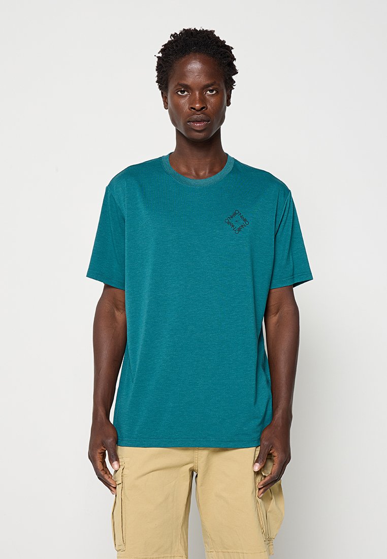 Mann mit kurzen lockigen Haaren trägt ein türkisfarbenes T-Shirt mit kleinem schwarzem Text auf der Brust und beige Cargoshorts steht vor weißem Hintergrund.