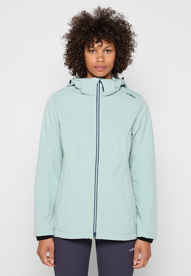 Frau mit lockigem Haar, die eine hellblaue Kapuzenjacke mit Reißverschluss und dunkle Hose trägt, steht vor einem schlichten hellen Hintergrund.