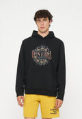 Junger Mann mit lockigem Haar, der einen schwarzen O'Neill Hoodie und gelbe Shorts mit schwarzem Text auf einfachem weißem Hintergrund trägt.