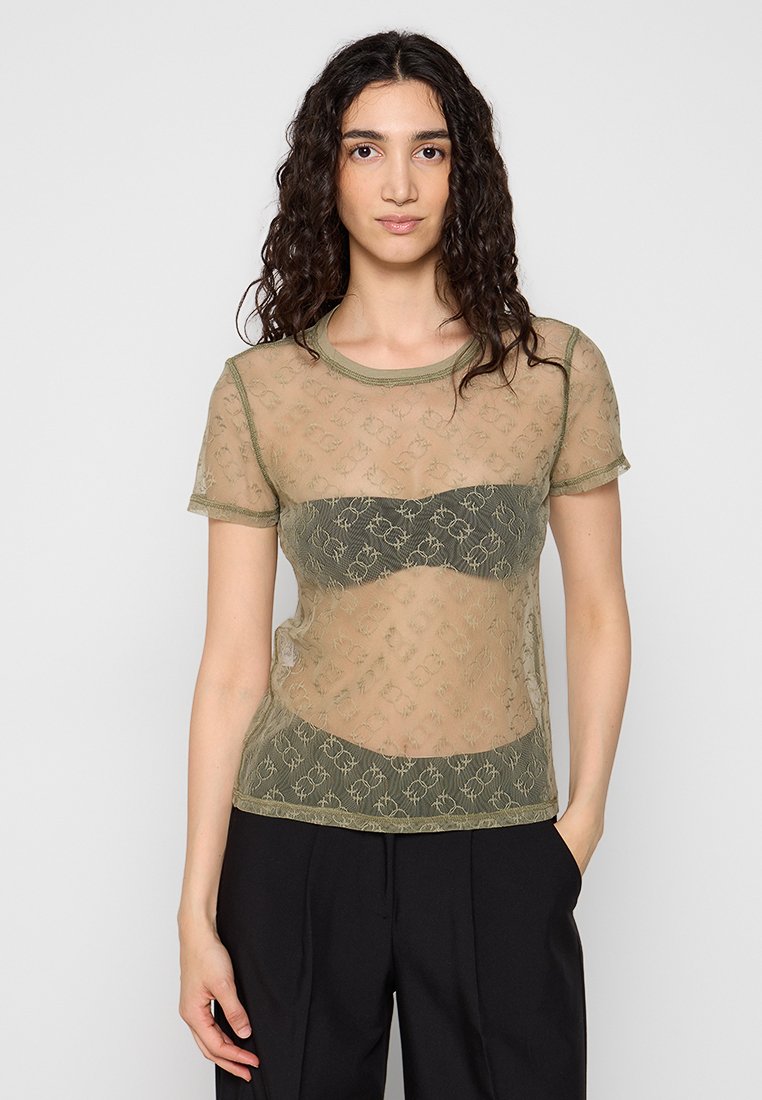 Frau mit lockigem Haar, die ein olivgrünes, durchsichtiges Kurzarm-Oberteil über einem passenden Bralette und schwarzer Hose trägt, und mit einer Hand in der Tasche steht.