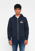 Junger Mann, der einen marineblauen Reißverschluss-Hoodie mit "DIESEL"-Logo auf der Brust trägt, kombiniert mit blauen Jeans, vor einem schlichten weißen Hintergrund stehend.