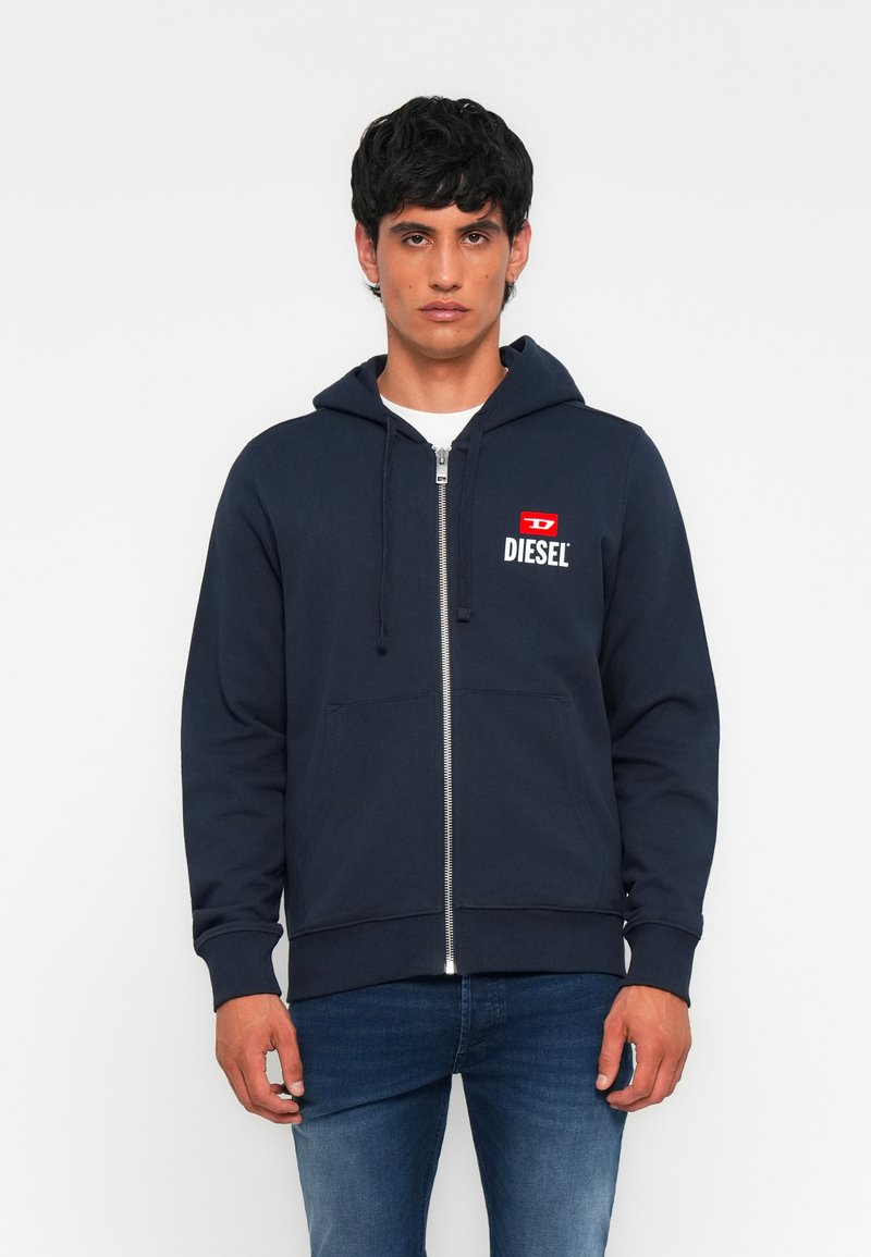 Junger Mann, der einen marineblauen Reißverschluss-Hoodie mit "DIESEL"-Logo auf der Brust trägt, kombiniert mit blauen Jeans, vor einem schlichten weißen Hintergrund stehend.