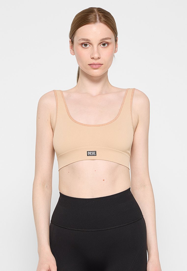 Beige gerippter, kurzer Bralette mit breitem Bund, der auf der Vorderseite ein schwarzes Logoetikett trägt, kombiniert mit schwarzen hochgeschnittenen Leggings.