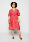 Frau lächelt, trägt ein knielanges rotes Kleid mit Blumenmuster und flatternden Ärmeln sowie rote Sandalen mit Absatz und Blumendetails.