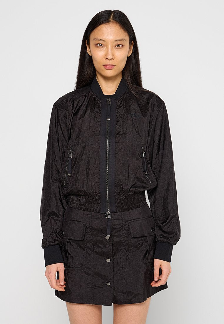 Schwarze Bomberjacke aus krinkligem Material, mit einem Reißverschluss vorne, zwei Reißverschlusstaschen und gerippten Bündchen. Kombiniert mit einem passenden Rock.