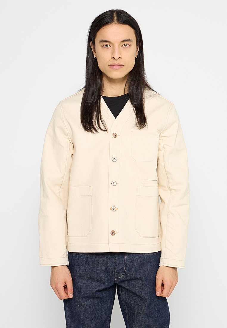 Beige Canvasjacke mit V-Ausschnitt, Knopfverschluss und zwei Fronttaschen. Besitzt eine strukturierte Oberfläche und braune Knöpfe als Kontrast.