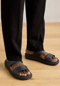 Schwarze Sandalen mit einer glatten, matten Oberfläche, die zwei breite Riemen über den Fuß und eine dicke, strukturierte Sohle aufweisen. Getragen mit schwarzen Hosen.