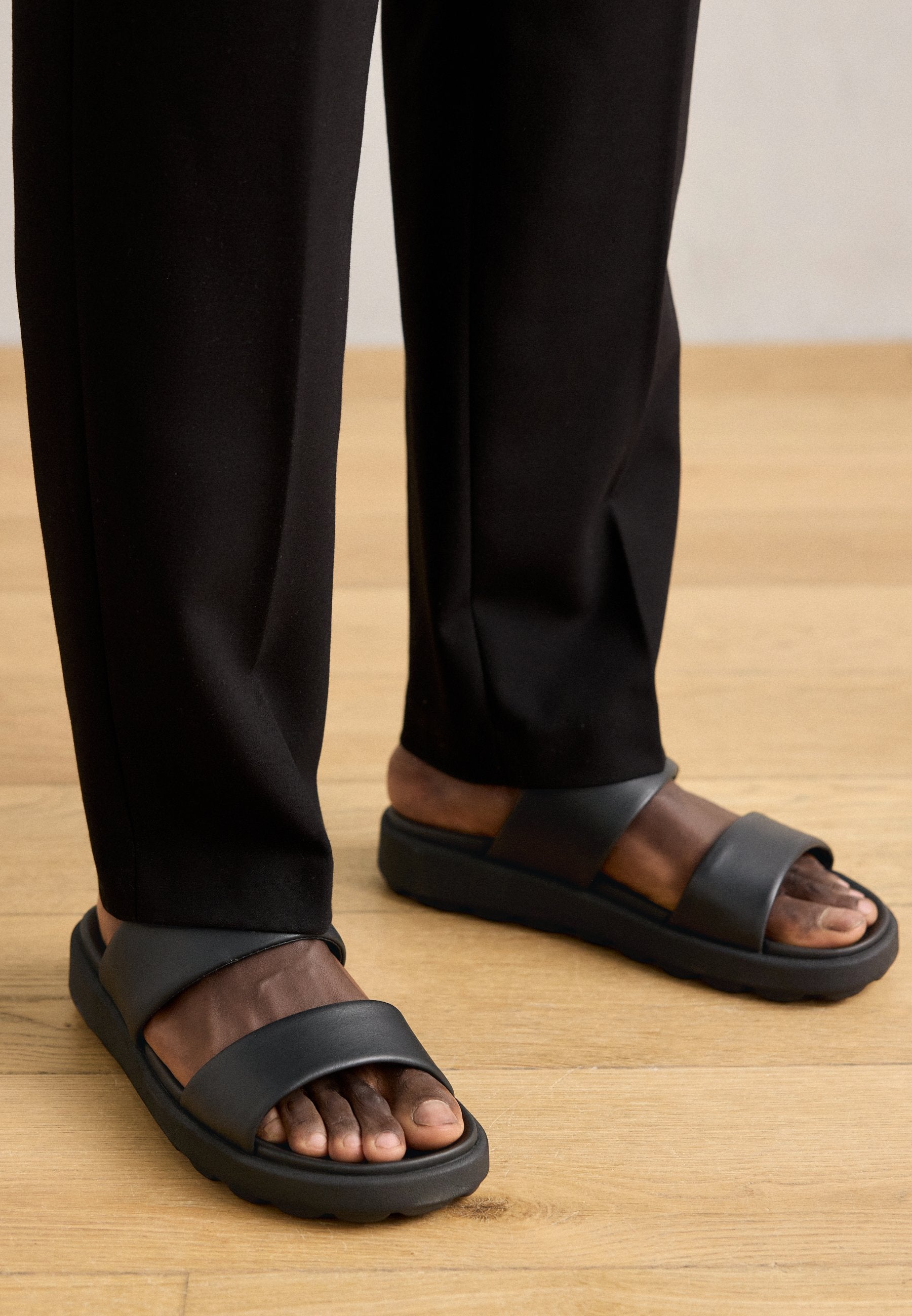 Schwarze Sandalen mit einer glatten, matten Oberfläche, die zwei breite Riemen über den Fuß und eine dicke, strukturierte Sohle aufweisen. Getragen mit schwarzen Hosen.