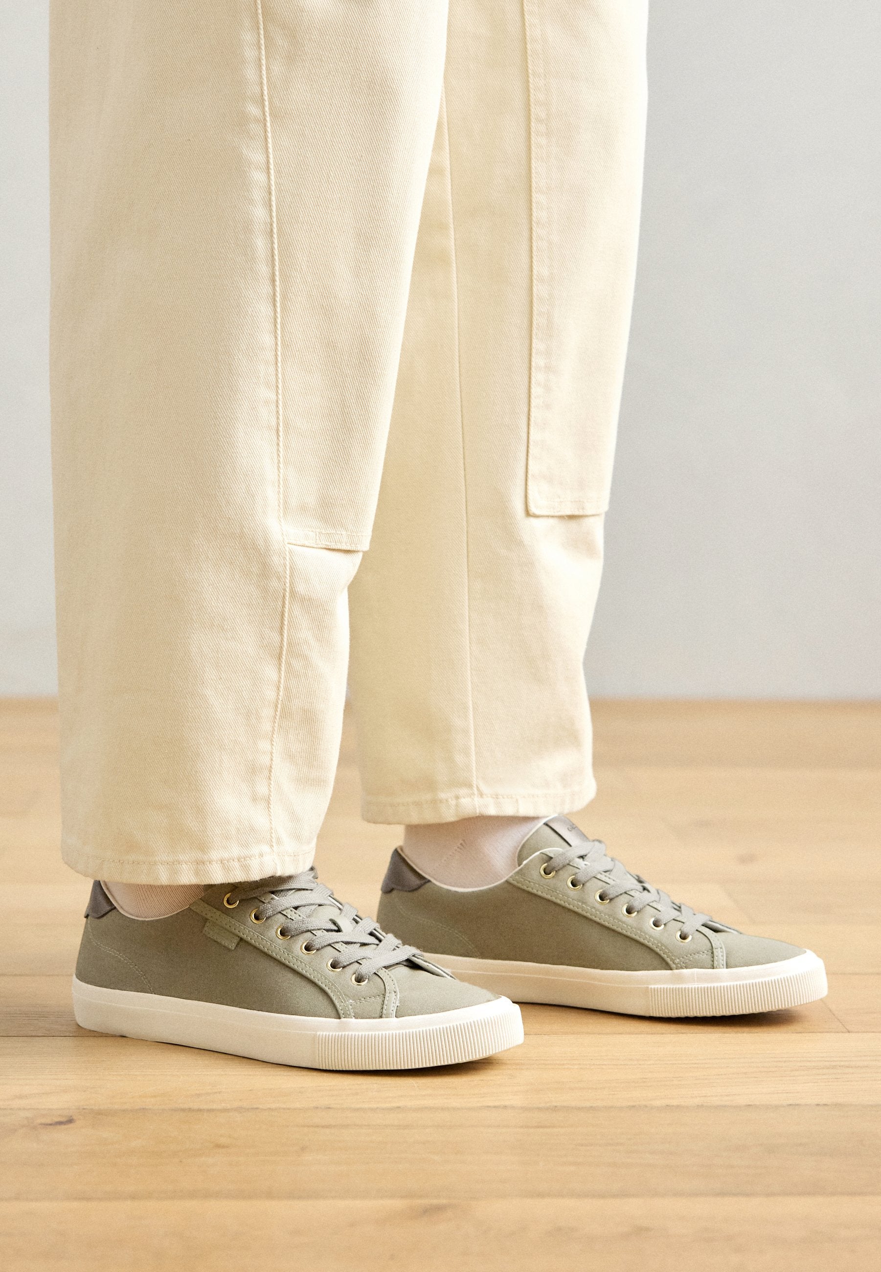 Hellgrüne Canvas-Sneakers mit grauen Akzenten, flachen Schnürsenkeln und einer strukturierten Gummisohle, kombiniert mit creamfarbenen, locker sitzenden Hosen.