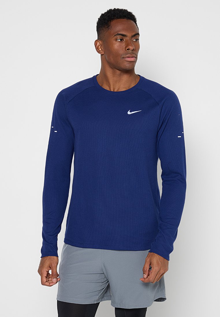 Athletischer Mann trägt ein dunkelblaues Nike Langarmshirt und graue Shorts, steht vor einem schlichten hellen Hintergrund und schaut zur Seite.