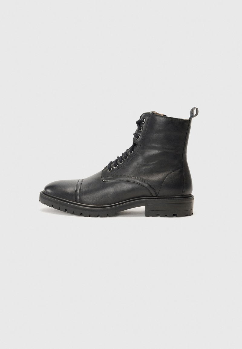 Schwarze Leder-Schnürstiefelette mit Kappenzehe, robuster Gummisohle und Zuglasche an der Rückseite auf einem schlichten hellen Hintergrund.