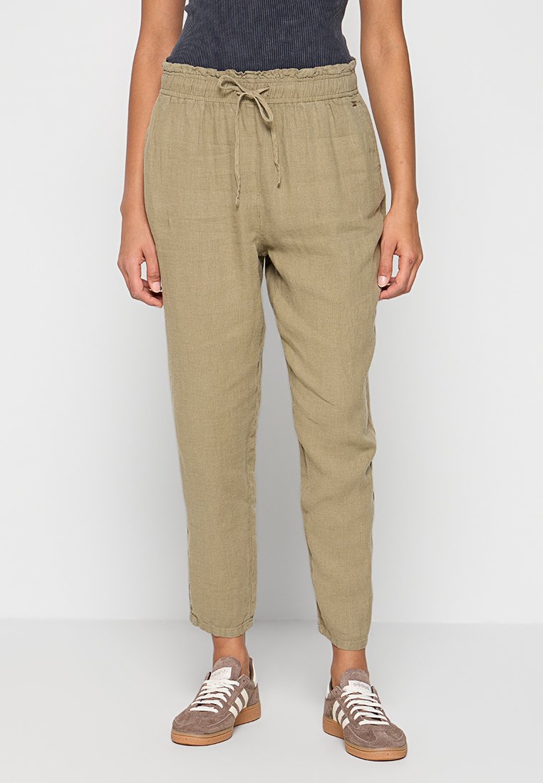 Olivgrüne Leinenhose mit elastischem Bund und Kordelzug, ausgestellten Beinen und strukturierter Oberfläche. Abgerundet durch braune Sneaker.