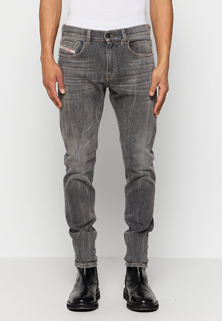 Mann in verblassten grauen Slim-Fit-Jeans, schwarzen Lederstiefeln und einem weißen ärmellosen Oberteil, der sich vor einem schlichten hellen Hintergrund aufhält.
