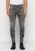 Mann in verblassten grauen Slim-Fit-Jeans, schwarzen Lederstiefeln und einem weißen ärmellosen Oberteil, der sich vor einem schlichten hellen Hintergrund aufhält.