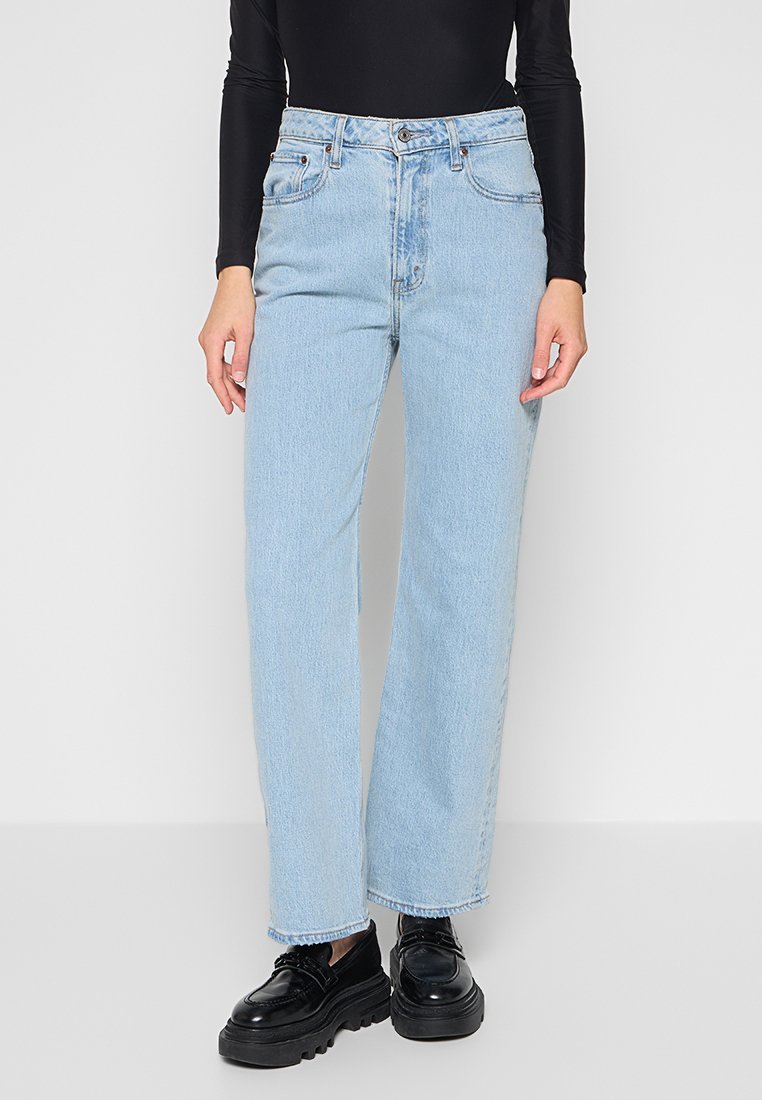 Hellblaue, hochtaillierte, weit geschnittene Jeans mit Knopfverschluss, fünf Taschen-Design und fransigem Saum. Getragen mit einem taillierten schwarzen Oberteil.