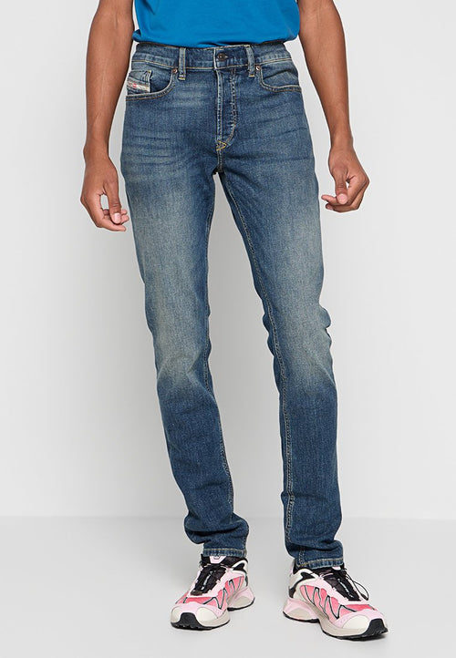 Mann in ausgewaschenen blauen Jeans, einem blauen Hemd und Sneakers in Pink, Weiß und Schwarz, der vor einem schlichten grauen Hintergrund steht.