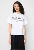Frau mit lockigem dunklem Haar, die ein weißes AllSaints-T-Shirt und einen schwarzen Spitzenrock trägt, vor einem schlichten hellen Hintergrund stehend.