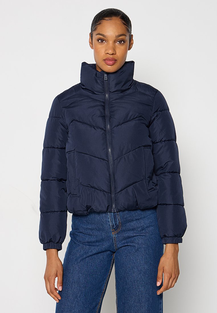 Marineblaue Steppjacke mit hohem Kragen, horizontalem Steppmuster, durchgehendem Reißverschluss und elastischen Bündchen; kombiniert mit blauen Jeans.