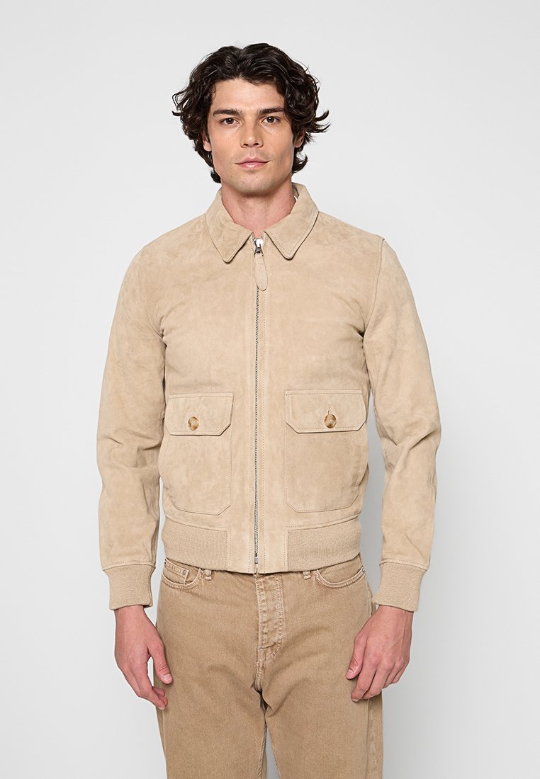Mann trägt eine beige Wildlederjacke mit Reißverschluss, Kragen und vorderen Klappentaschen, kombiniert mit beigen Hosen, steht vor einem schlichten Hintergrund.