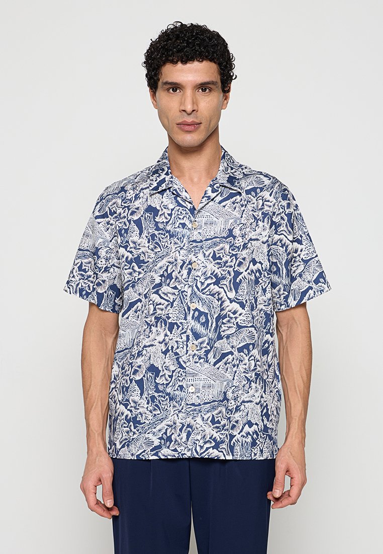 Mann trägt ein blau-weißes Hemd mit kurzem Ärmel und Blumen- sowie Tiermuster und dunkle Hose, steht vor einem einfarbigen Hintergrund.