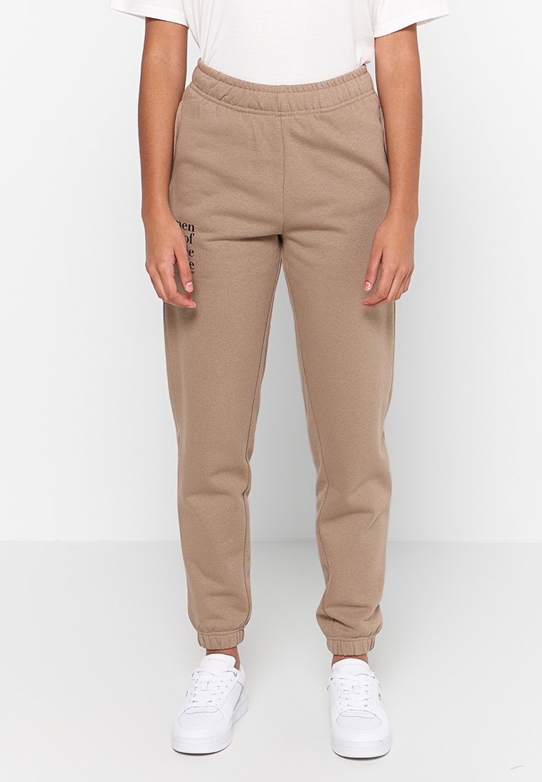 Beige Jogginghose mit elastischem Bund und gerippten Bündchen an den Knöcheln. Mit einem bedruckten Textdetail am linken Bein. Kombiniert mit weißen Sneakers.