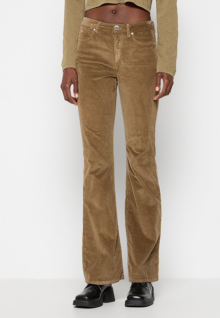 Person trägt braune Corduroy Schlaghosen, ein beigefarbenes, kurzes Langarmoberteil und schwarze Plateaustiefel vor einem einfarbigen Hintergrund.