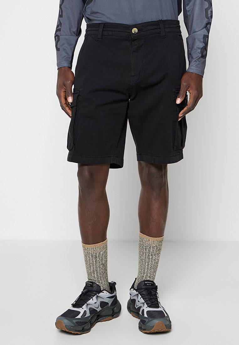 Mann trägt schwarze Cargo-Shorts, graues langärmeliges Shirt, beige Knöchelsocken und schwarze sowie graue Sportschuhe, steht vor einem schlichten Hintergrund.