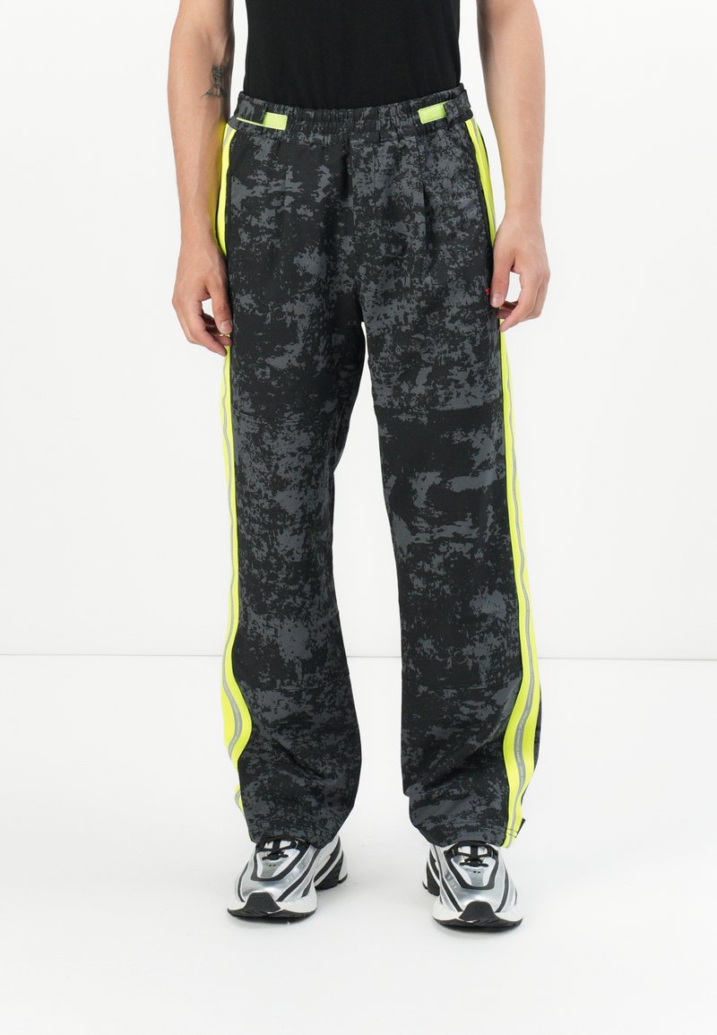 Schwarze Sweatpants mit neon-gelben Seitenstreifen, elastischem Bund und schmal zulaufenden Beinen, kombiniert mit grau-schwarzen Sneakers.