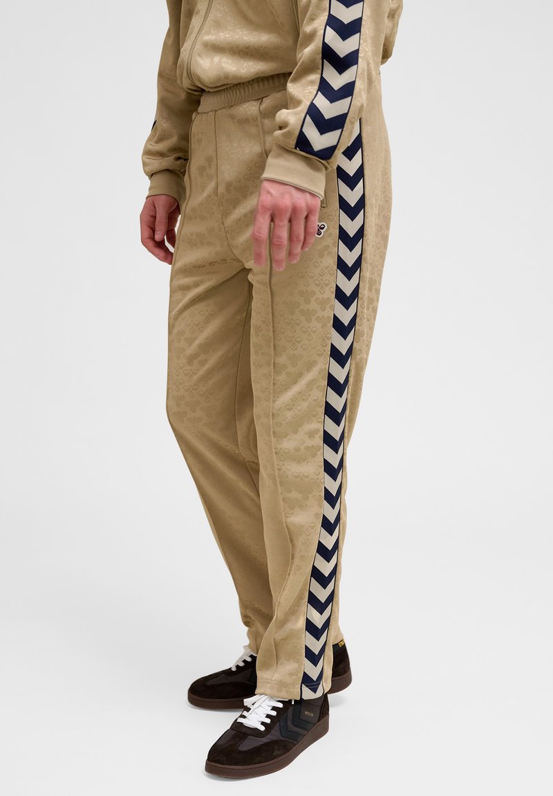 Beige Trainingshose mit dezentem Mickey-Mouse-Muster und marineblauen Chevron-Seitenstreifen, kombiniert mit schwarzen Wildledersneakern mit weißen Schnürsenkeln.