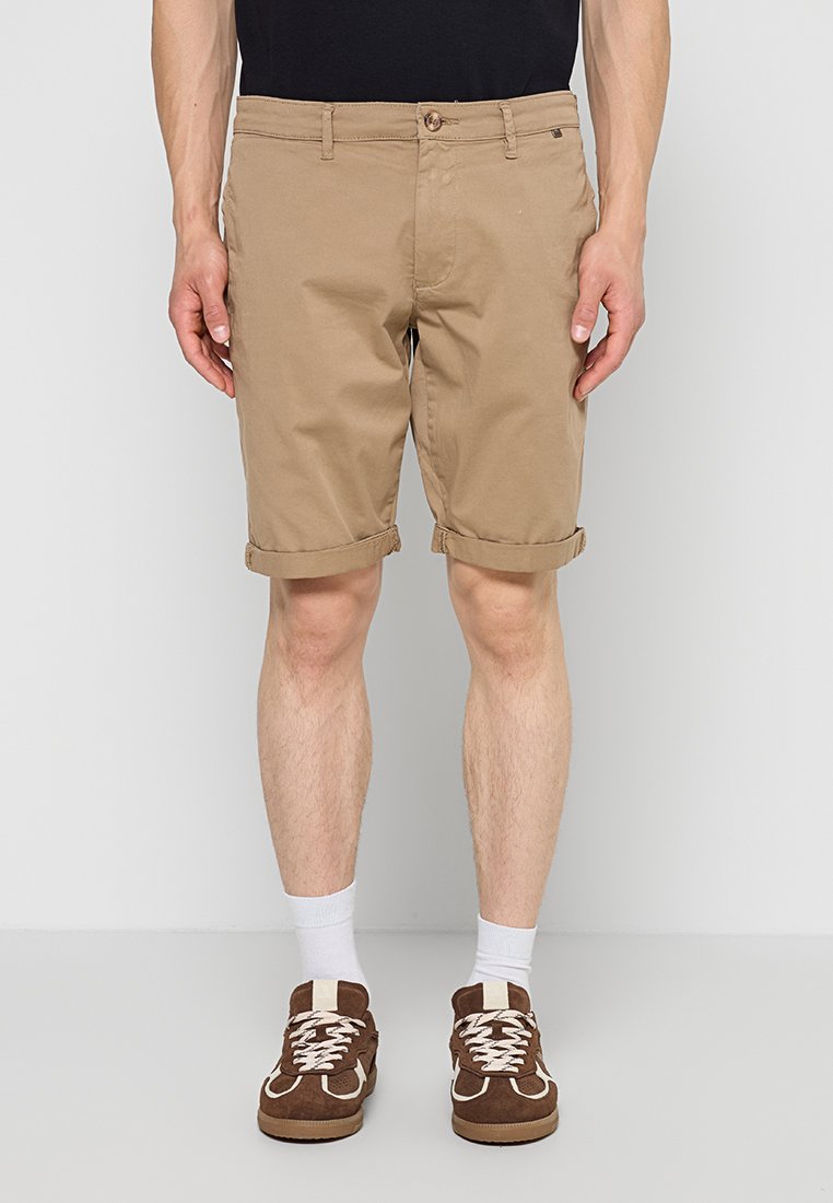 Mann trägt beige hochgekrempelte Shorts, weiße Socken und braune Turnschuhe und steht vor einem schlichten hellen Hintergrund.