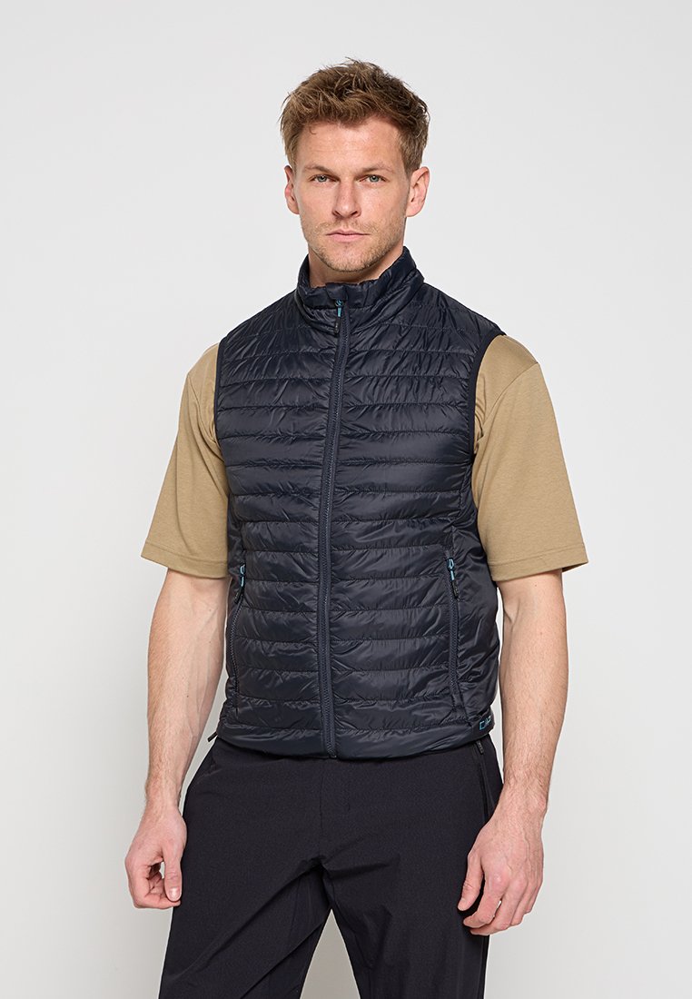 Mann trägt eine schwarze gesteppte ärmellose Weste über einem beigen kurzärmligen Hemd und schwarzen Hosen, steht vor einem schlichten hellen Hintergrund.
