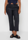 Person, der schwarze schmal gestreifte, cropped Hosen, eine dunkelblaue Weste und schwarze Plateausandalen ohne Riemen trägt, vor einem schlichten weißen Hintergrund.