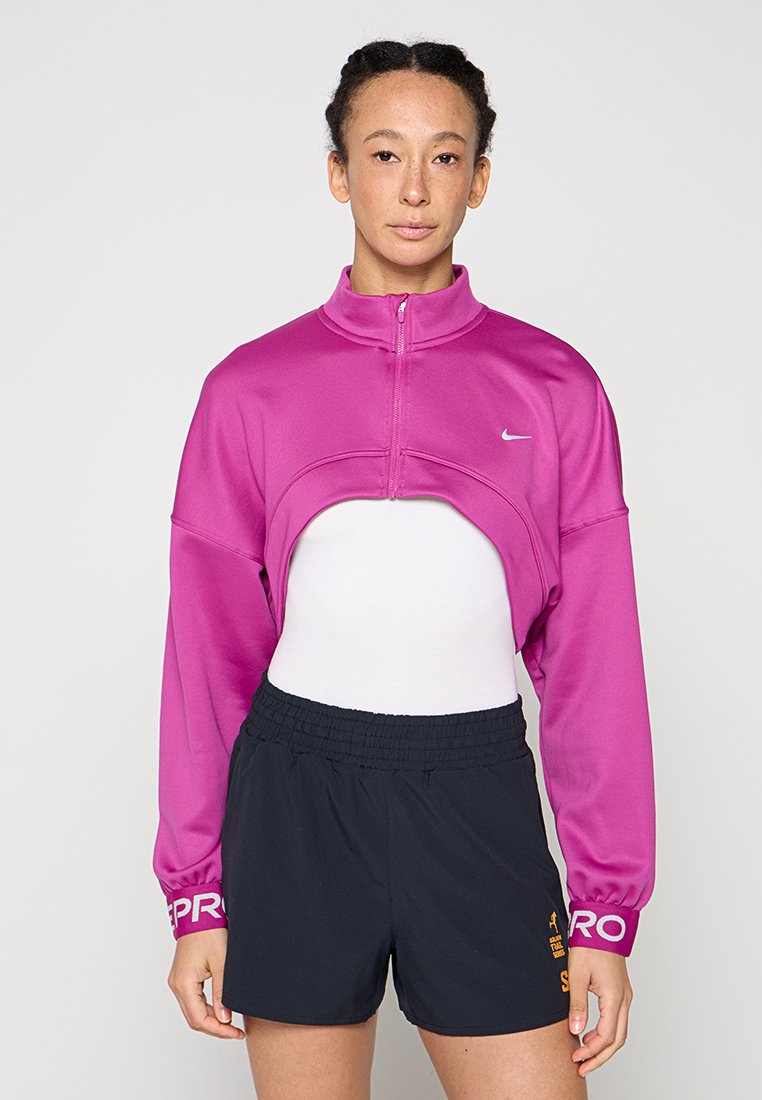 Frau trägt eine kurze magentafarbene Jacke mit Reißverschluss über einem weißen Oberteil, kombiniert mit schwarzen elastischen Sportshorts vor einfarbigem Hintergrund.