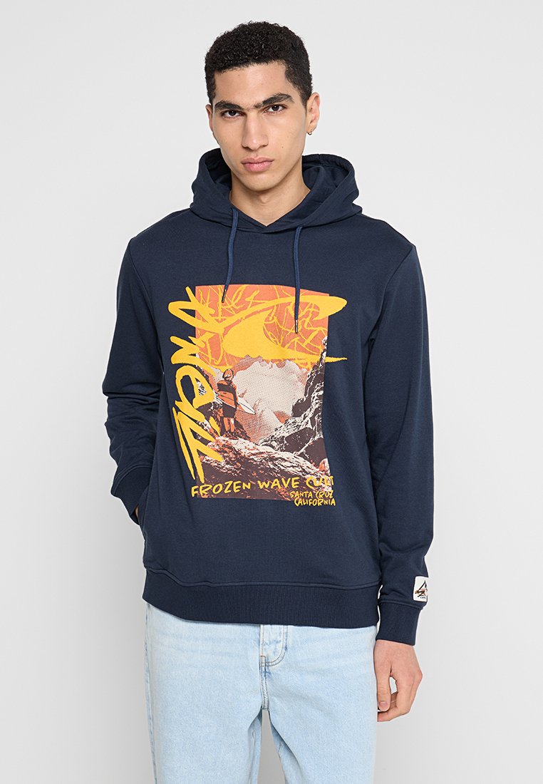 Junger Mann trägt einen dunkelblauen Hoodie mit gelbem und orangefarbenem Surf-Motiv und hellblauen Jeans und steht vor einem einfarbigen grauen Hintergrund.