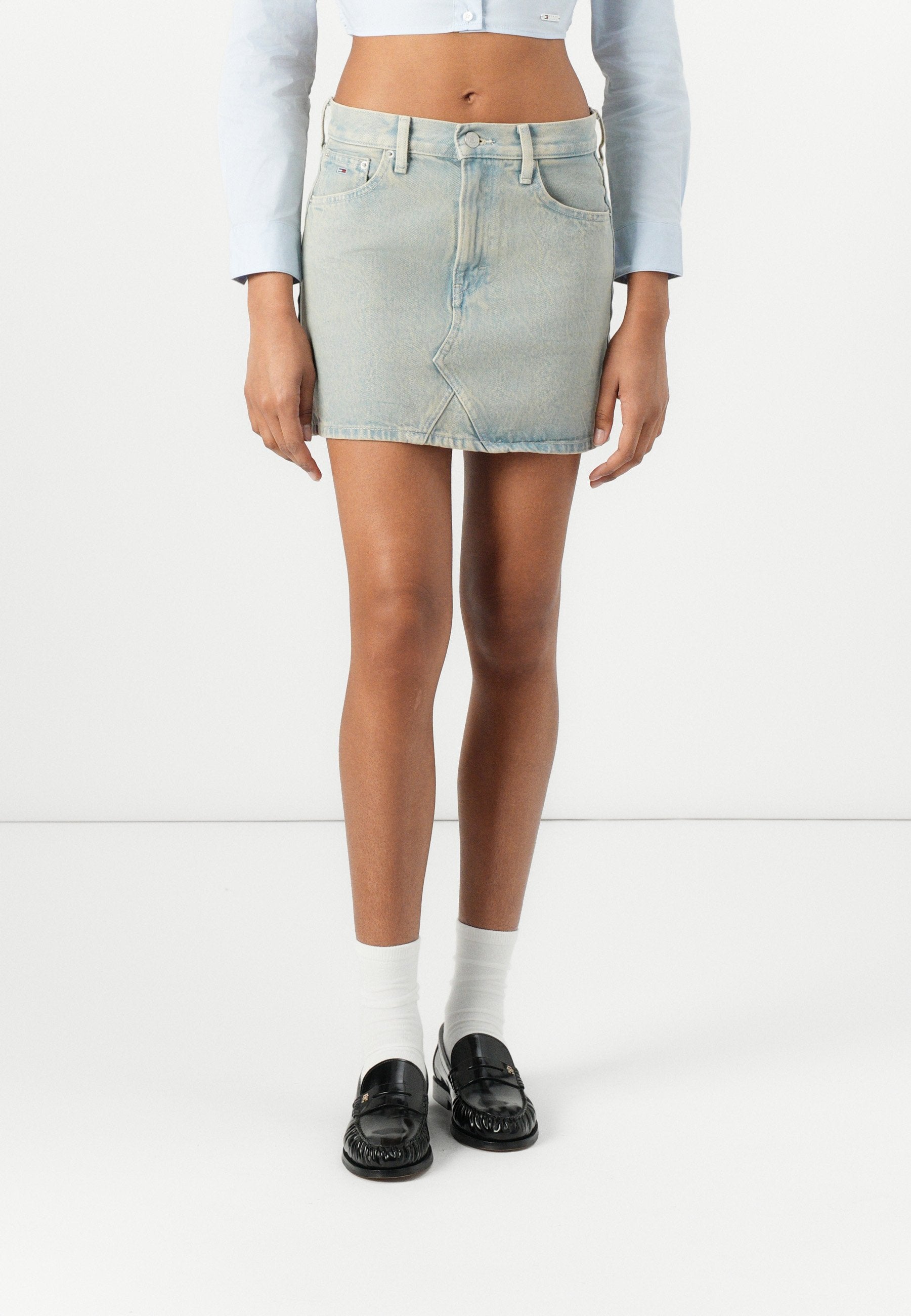 Hellblaue Denim-Minirock mit geradem Schnitt, ausgestattet mit Vordertaschen und einem dezenten ausgeblichenen Muster. Kombiniert mit schwarzen Slippern und weißen Socken.
