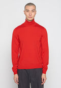 Mann mit kurzem Haar, der einen enganliegenden roten Rollkragenpullover und dunkelgraue Hosen trägt, steht vor einem einfarbigen hellen Hintergrund.