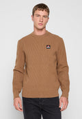 Junger Mann in einem braunen gerippten Pullover mit Diesel-Logo auf der Brust und schwarzen Jeans, der vor einem schlichten, hellen Hintergrund steht.