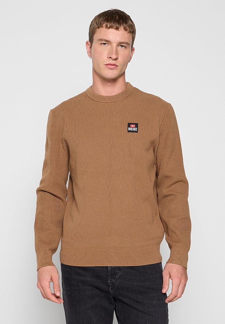 Junger Mann in einem braunen gerippten Pullover mit Diesel-Logo auf der Brust und schwarzen Jeans, der vor einem schlichten, hellen Hintergrund steht.