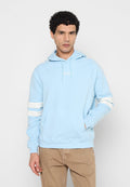 Mann trägt hellblauen Hoodie mit weißen Streifen an den Ärmeln und beigen Hosen, steht vor einem schlichten hellgrauen Hintergrund.