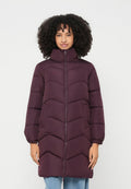 Frau mit lockigem Haar, die eine lange, gesteppte lila Daunenjacke mit hohem Kragen und seitlichen Taschen trägt und vor einem schlichten Hintergrund steht.