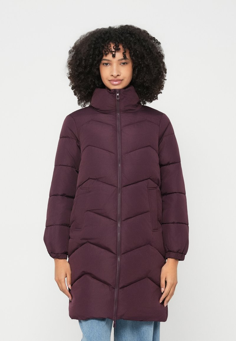 Frau mit lockigem Haar, die eine lange, gesteppte lila Daunenjacke mit hohem Kragen und seitlichen Taschen trägt und vor einem schlichten Hintergrund steht.