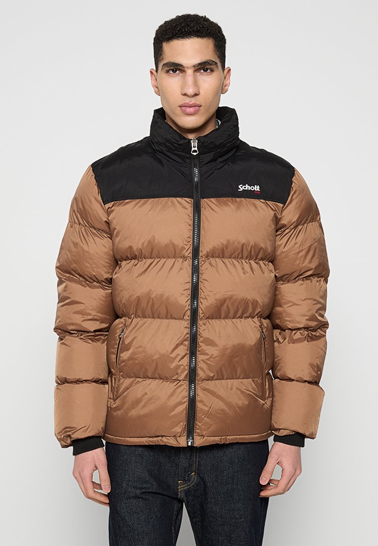 Mann, der eine braune und schwarze gepolsterte Jacke mit Reißverschlusstaschen trägt und vor einem schlichten hellen Hintergrund steht.