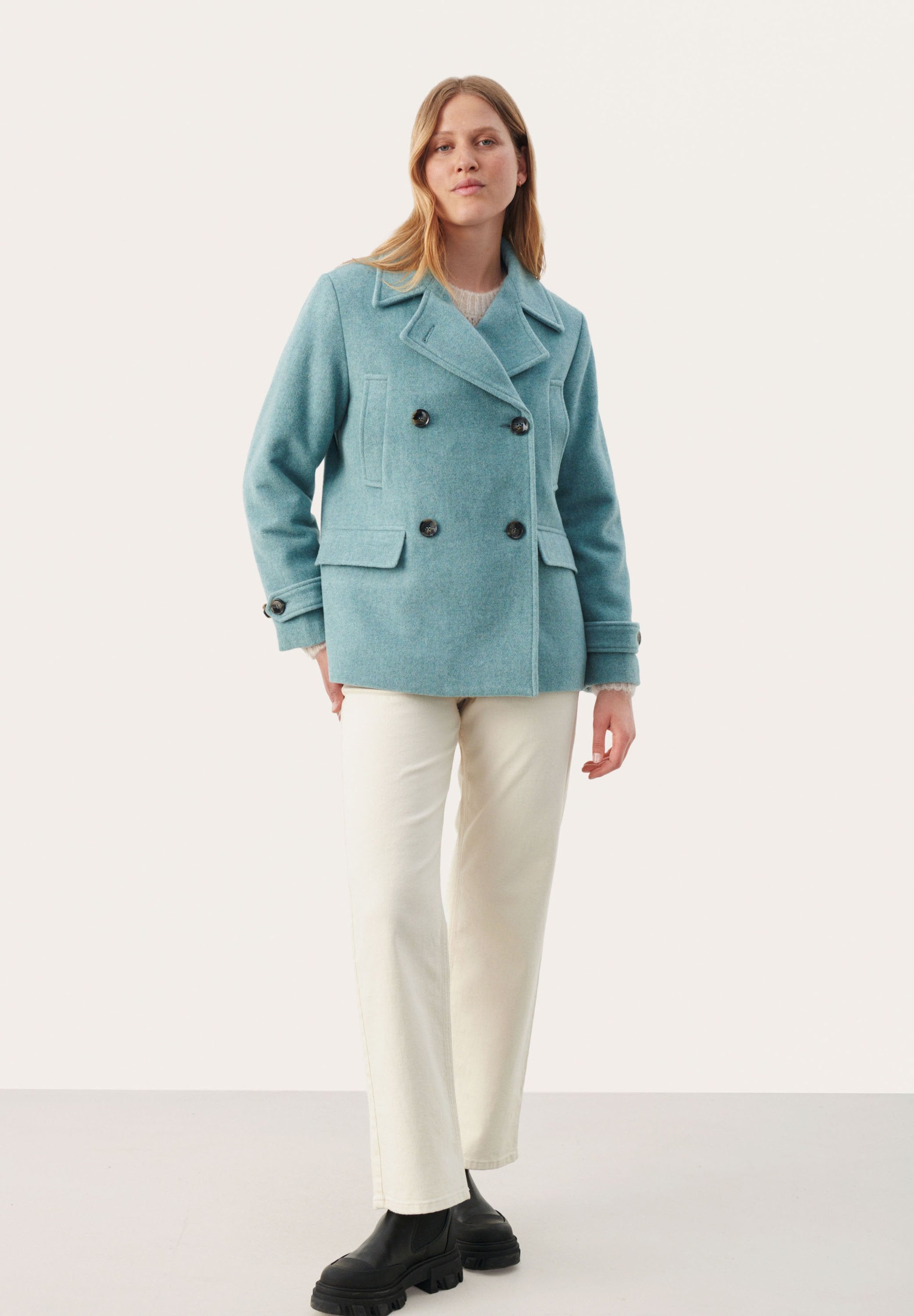 Hellblauer Woll-PEACOAT mit Doppelreihe, schwarzen Knöpfen und seitlichen Taschen, kombiniert mit cremefarbenen Schlaghosen und schwarzen Stiefeln.