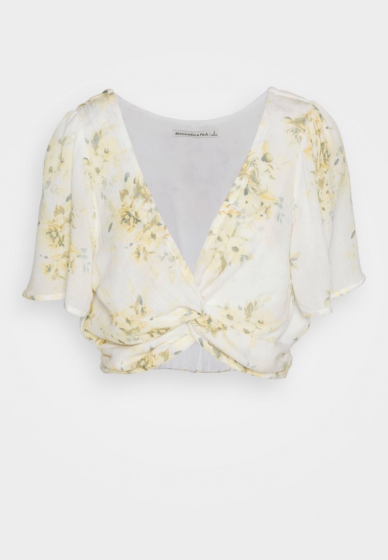 Blumenbluse mit Wickeldesign, kurzen Flatterärmel und verdrehtem Vorderteil. Die Farben bestehen aus zartem Gelb und Weiß mit einer dezenten Textur.