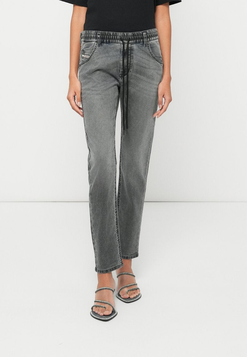 Eine Frau trägt graue, gewaschene Denim-Jogginghosen mit schwarzem Kordelzug und offenen Sandalen mit Riemchen und steht vor einem schlichten weißen Hintergrund.
